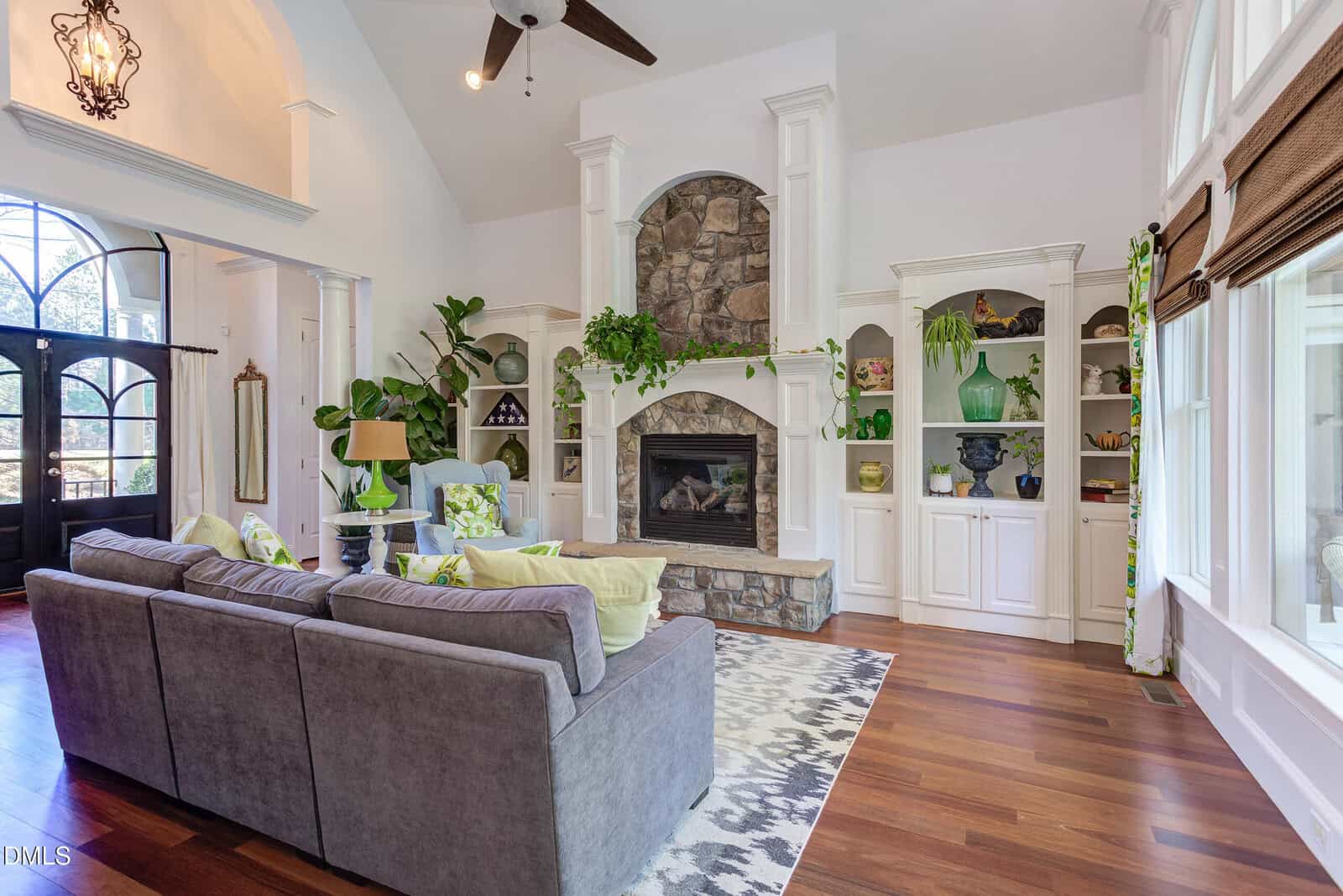 A living room with a stone fireplace, gray sectional sofa, patterned rug, built-in white shelves with decor, wood floors, large windows with blinds, and green plants throughout the space.