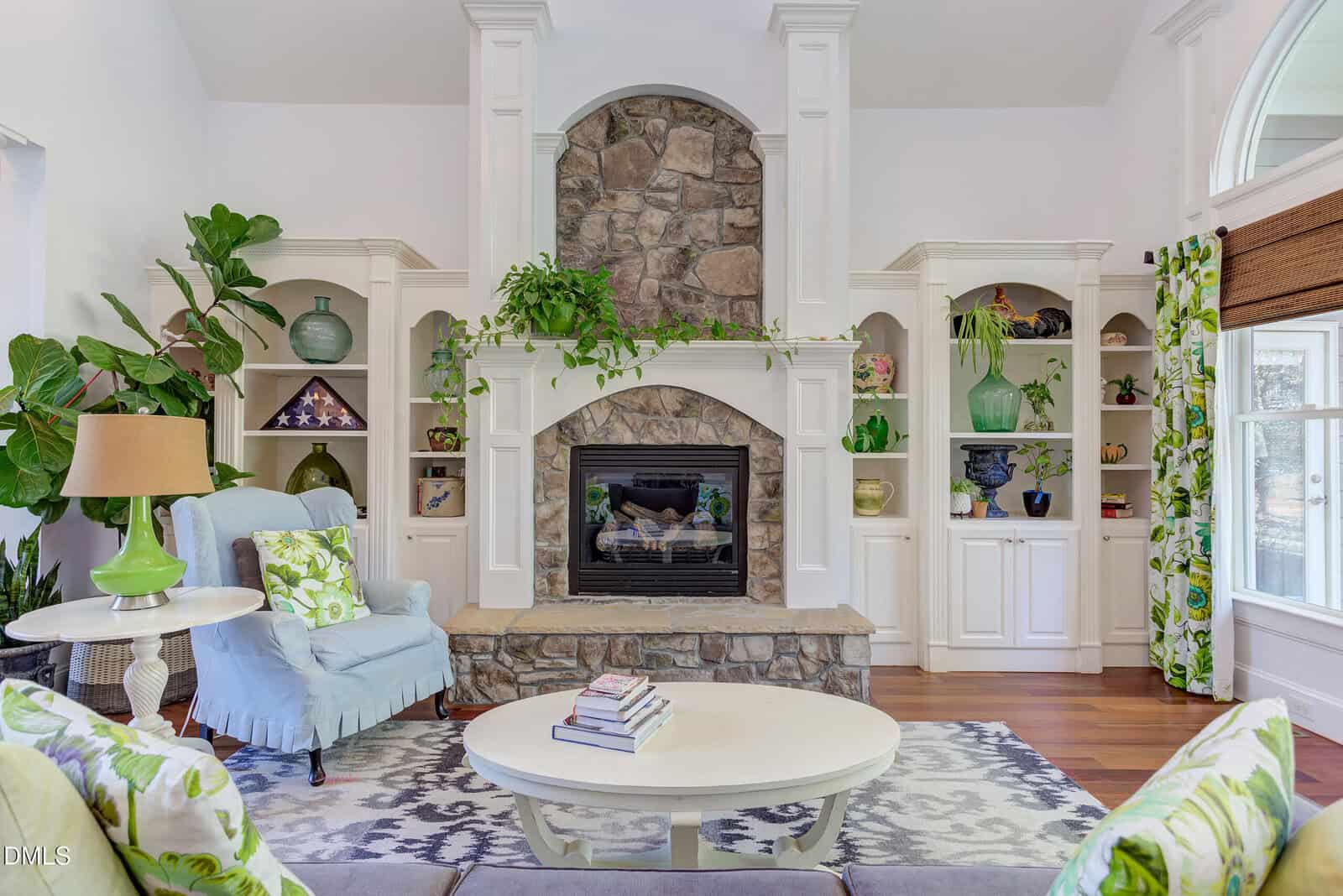 A living room with a stone fireplace, white built-in shelves with decorative items, green plants, a light blue armchair, a round white coffee table with books, and patterned curtains and cushions.