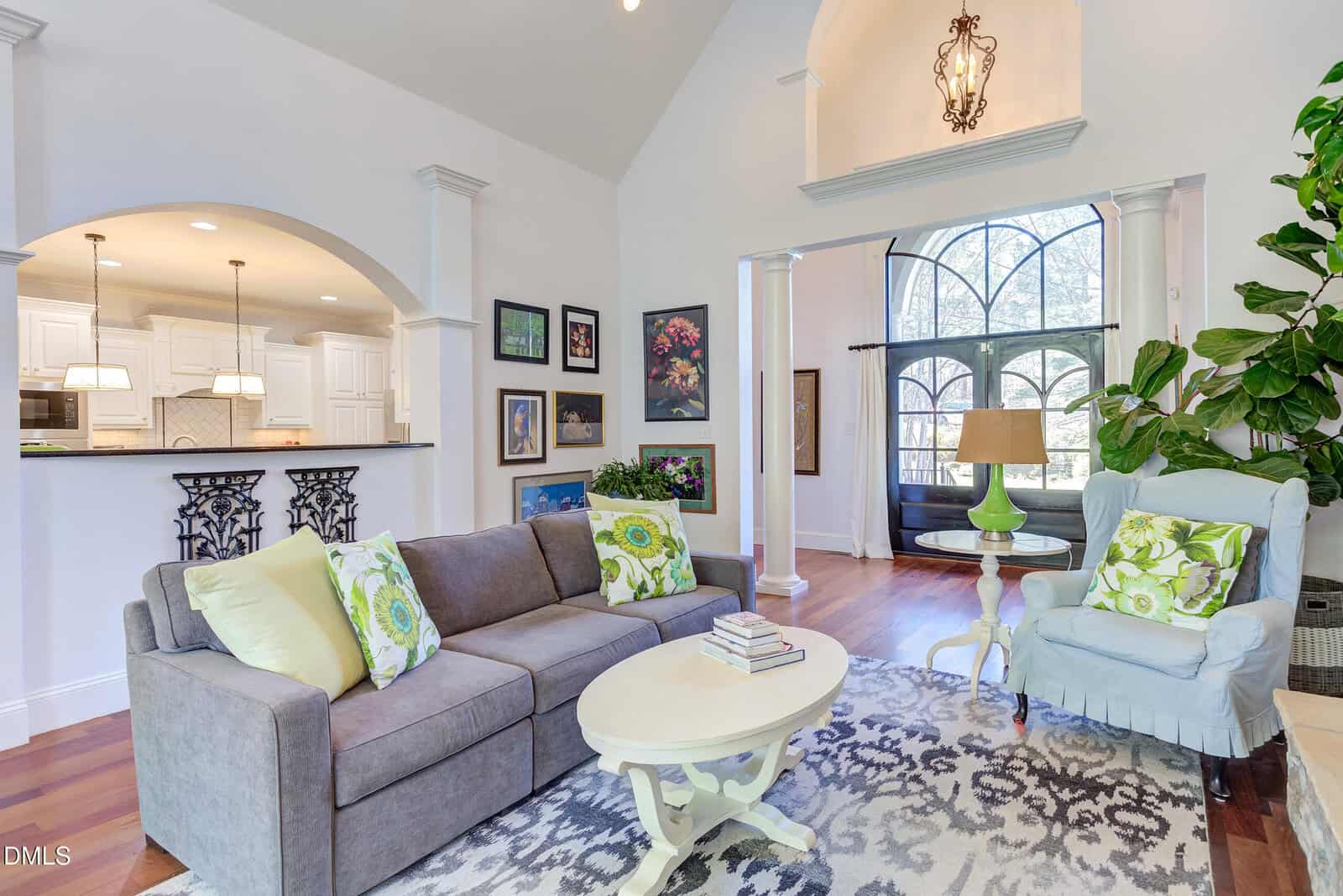 Bright living room with a gray sofa, green pillows, and a patterned rug. There’s an armchair, coffee table, wall art, large arched windows, and a view into a white kitchen with pendant lights.