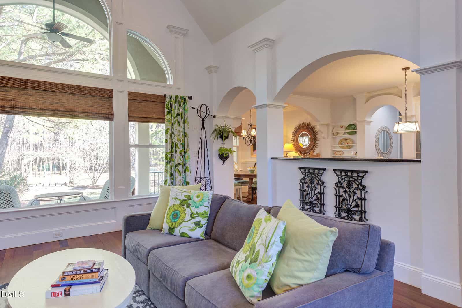 Bright living room with a gray sofa, green floral pillows, and large windows. There’s a round table with books, arched doorways leading to a dining area, and sunlight streaming in from outside.