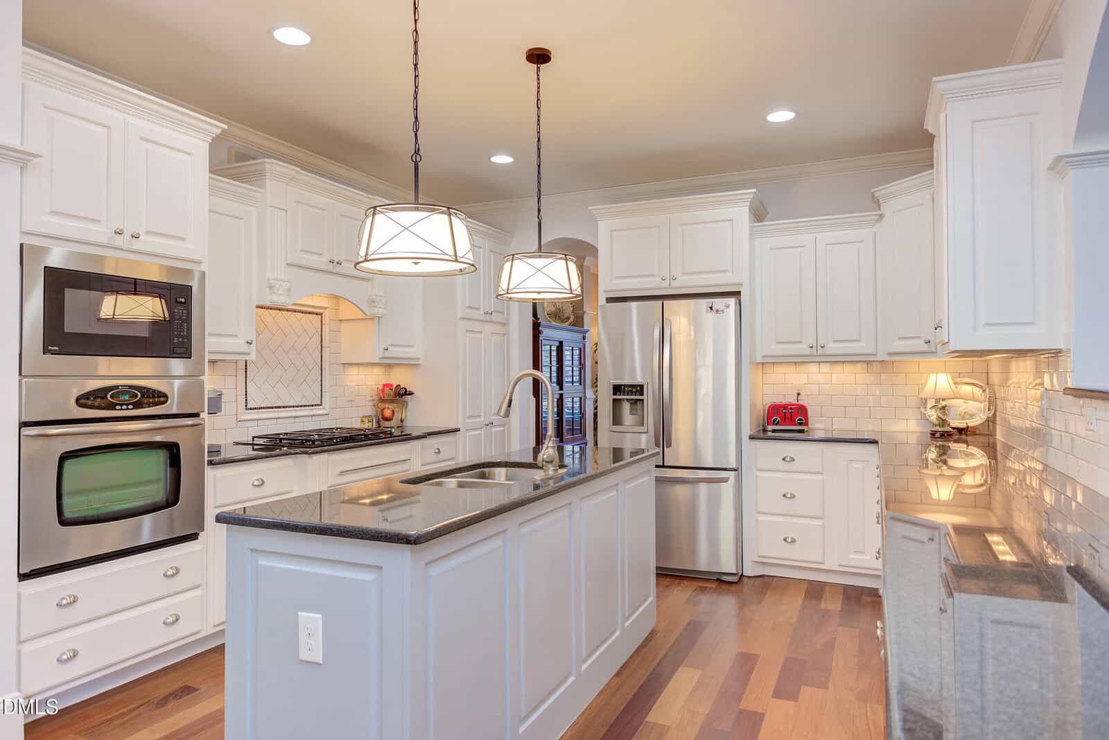 Modern kitchen with white cabinets, stainless steel appliances, black countertops, a central island with a sink, pendant lights, wood floors, and a red toaster on the counter.