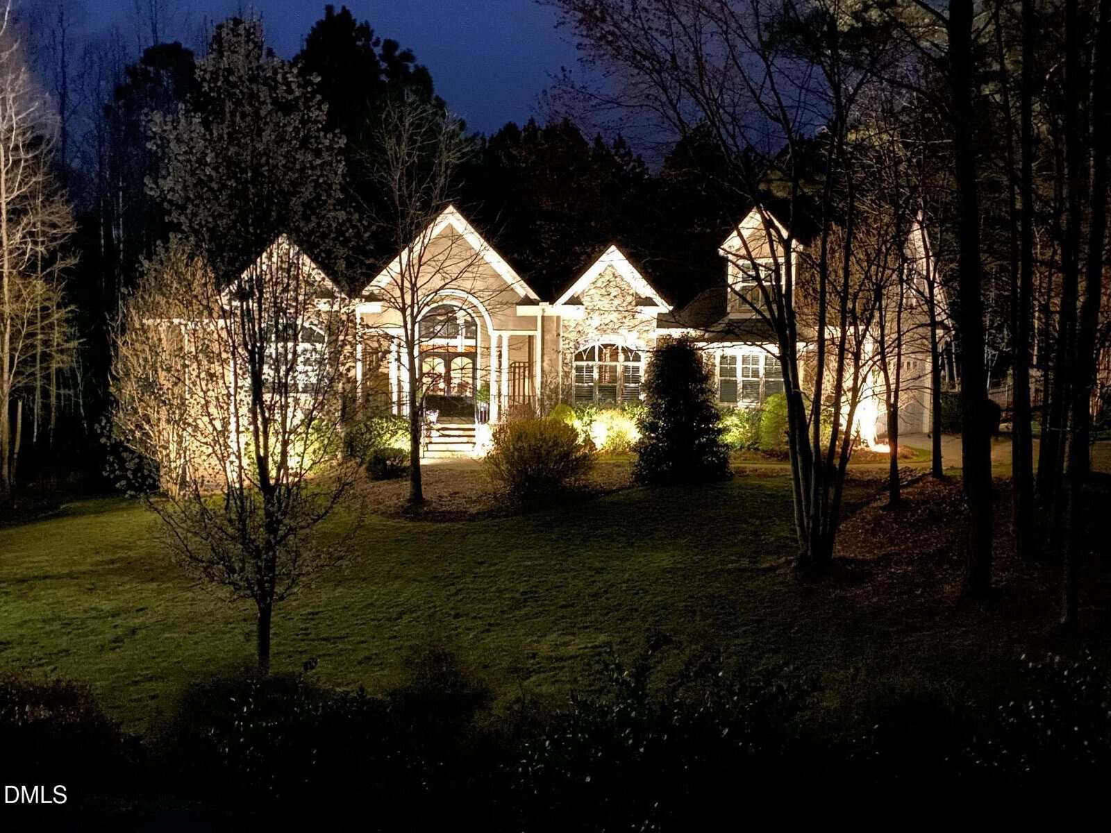 A single-story house with large front windows is illuminated at night by exterior lights, surrounded by trees and a well-maintained lawn. The sky is dark, and the yard features several shrubs and small trees.