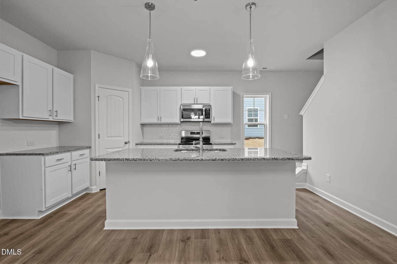 Modern kitchen with white cabinets, granite countertops, stainless steel appliances, pendant lights over a large island, and wood-look flooring. A door and stairs are visible in the background. Room has a clean, minimalist design.