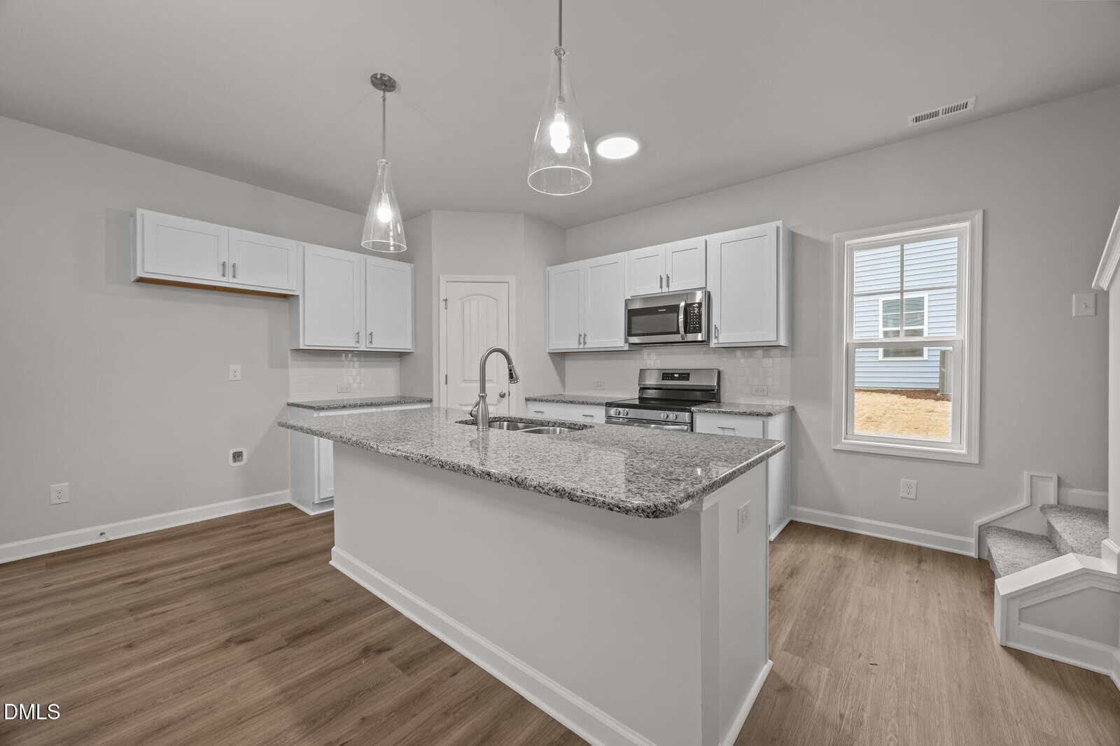 Modern kitchen with white cabinets, stainless steel appliances, granite countertops, pendant lights, and wood flooring. A large window provides natural light, and stairs are visible on the right side.