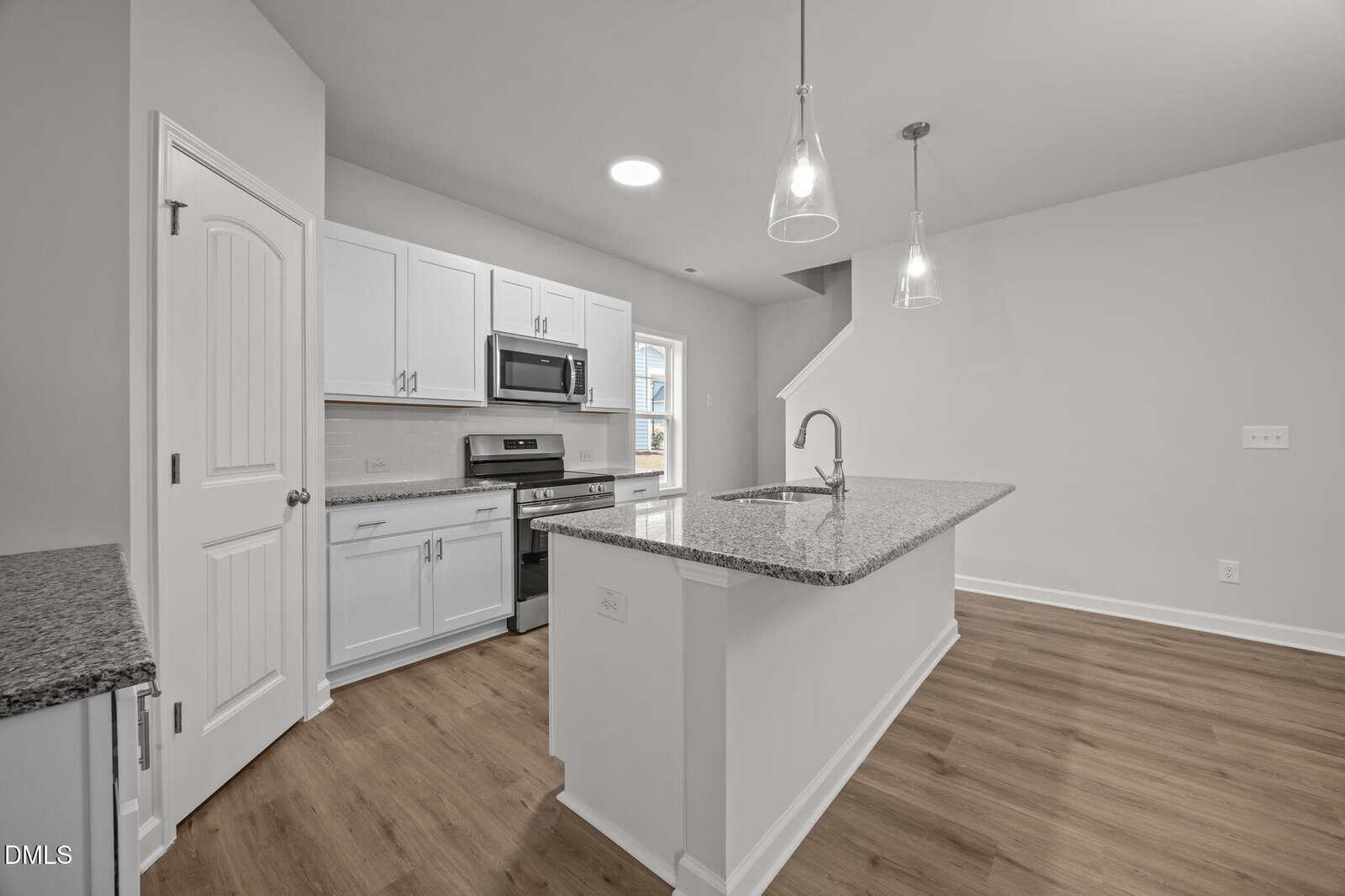 Modern kitchen with white cabinets, stainless steel appliances, granite countertops, and an island with pendant lights. Light wood flooring and neutral walls create a bright, open space. Stairs visible in the background.