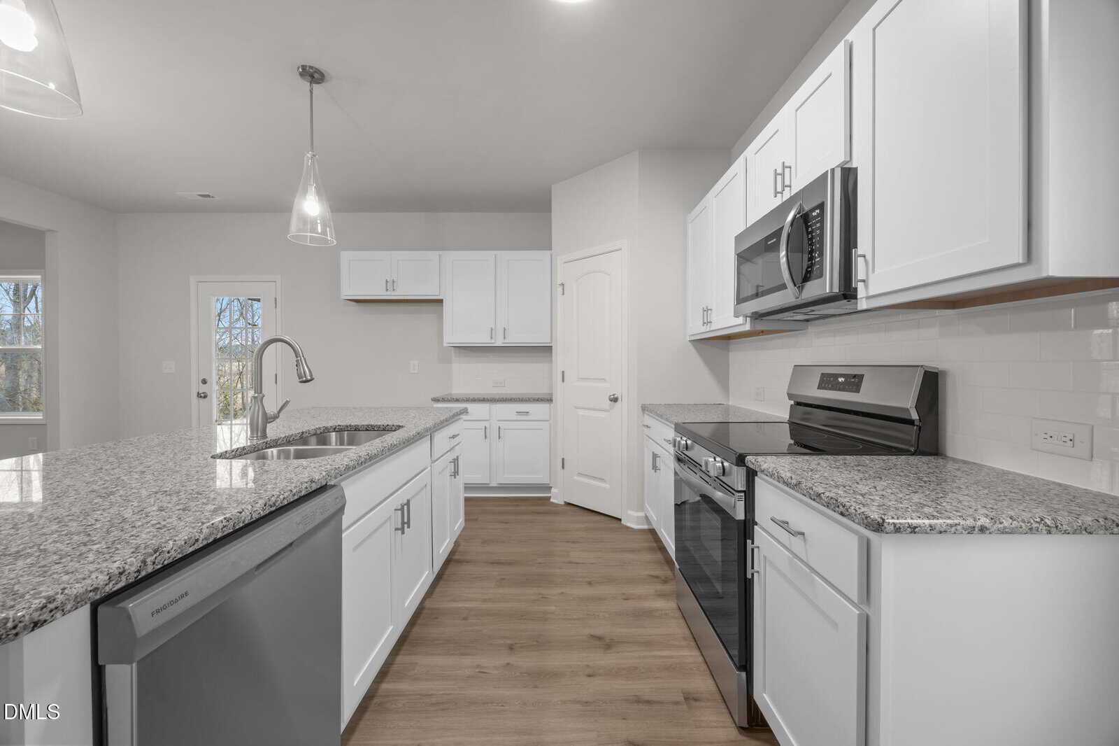 Modern kitchen with white cabinets, granite countertops, stainless steel appliances, sink on island, wood flooring, pendant lighting, and a pantry door. A window and exterior door are visible in the background.