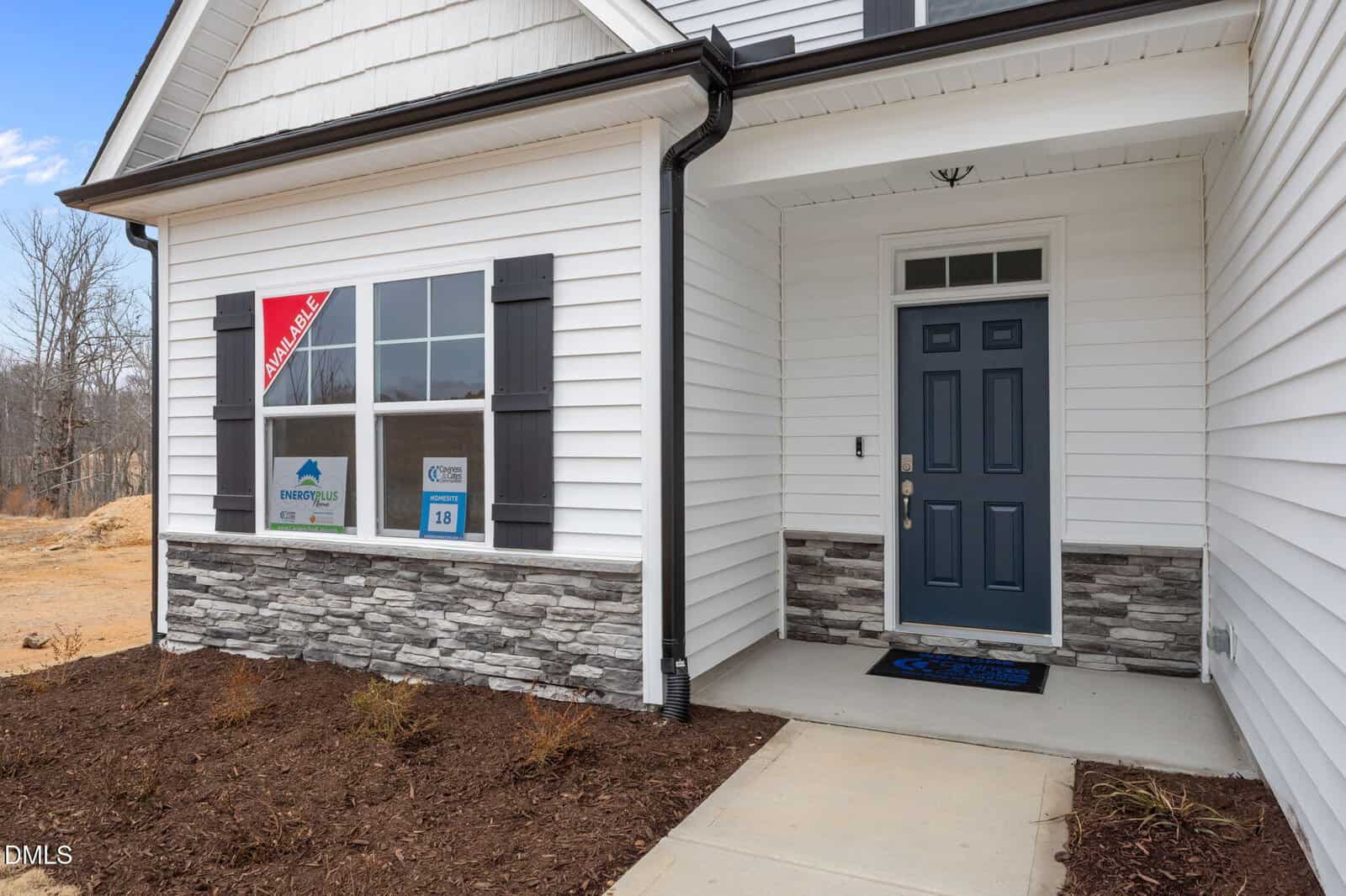 Front entrance of a modern house with white siding, dark shutters, and stone accents. The door is dark blue with a welcome mat. Mulch and soil border the walkway; window displays temporary signs.