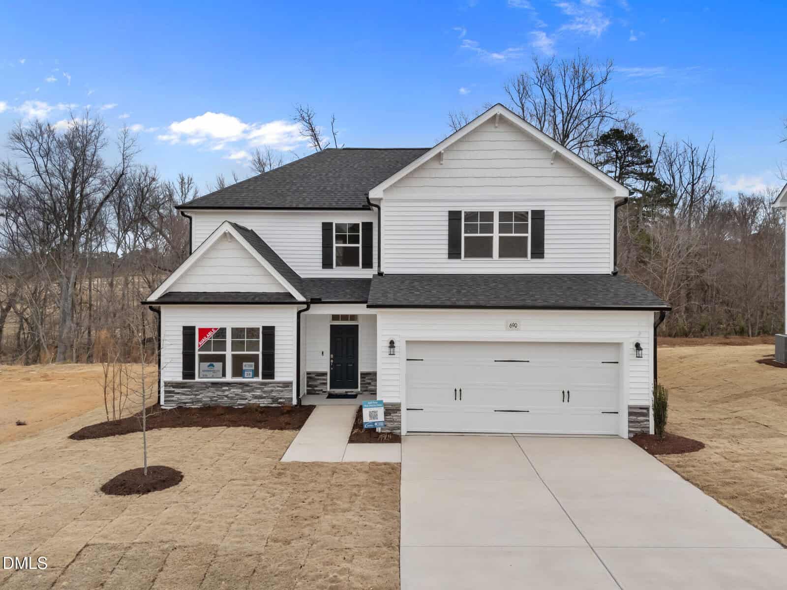 Two-story white house with a double garage, black roof, and minimal landscaping. The front yard has brown grass and a straight driveway. The house is set against a backdrop of leafless trees under a blue sky with a few clouds.
