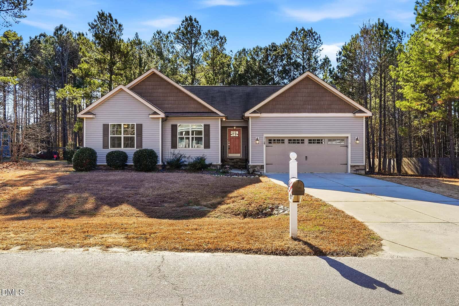 Charming single-story house at 240 N Ridge Drive, Louisburg, with beige siding, brown gables, and a two-car garage. Concrete driveway and mailbox in front, plus a lawn and trees under a clear blue sky.