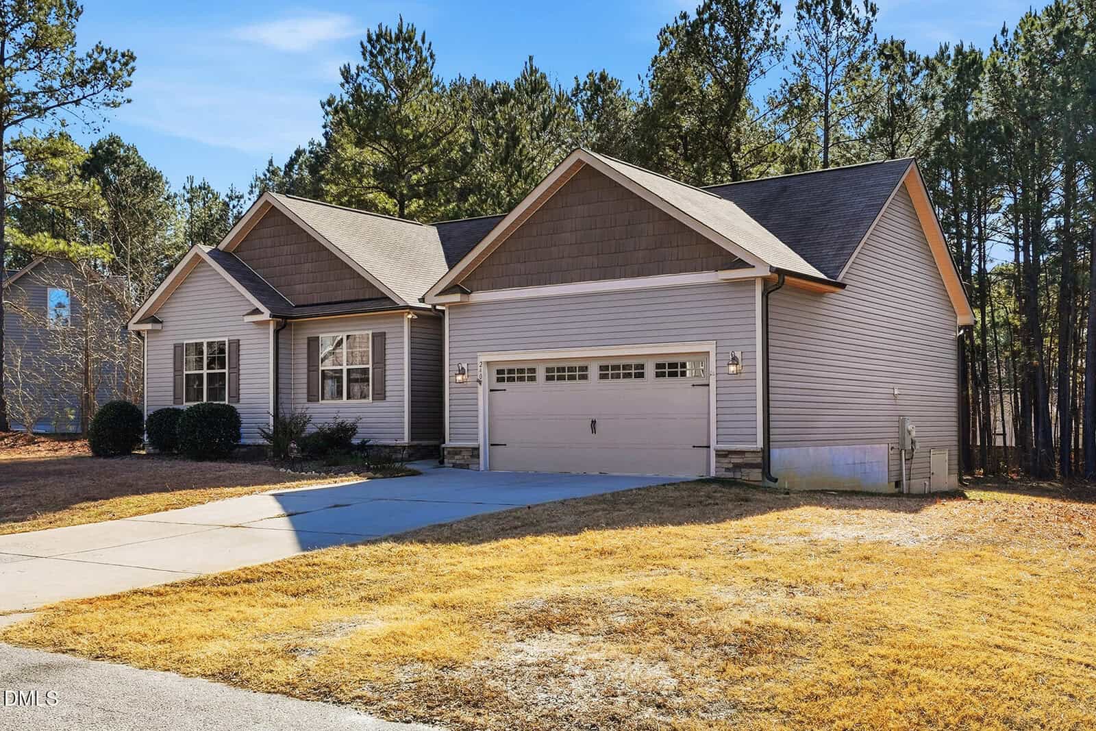 Single-story suburban house with gray siding, a gabled roof, and a two-car garage. The front yard has a concrete driveway and patchy grass. Trees and other houses are visible in the background.
