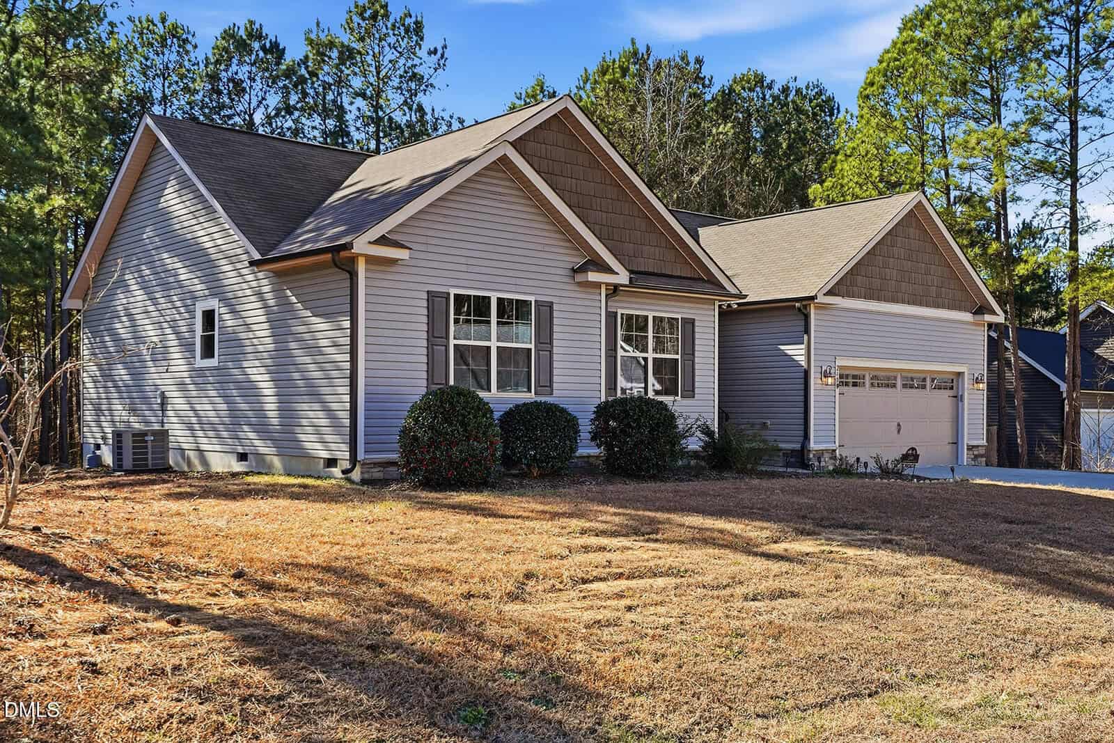 Single-story house with gray siding, two front-facing gables, white trim, and a double garage. The yard has dry grass and several bushes, and pine trees are in the background under a clear sky.