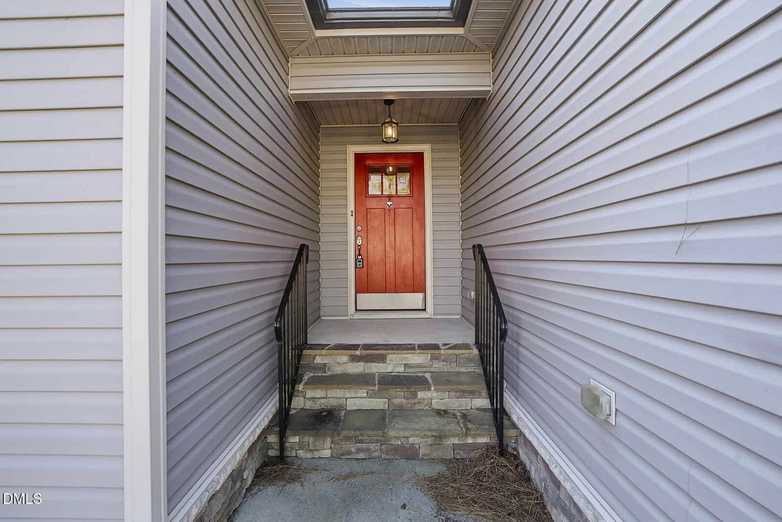 A straight-on view of a red front door with a window, located at the end of a covered entryway with gray siding, stone steps, and black metal railings on both sides.