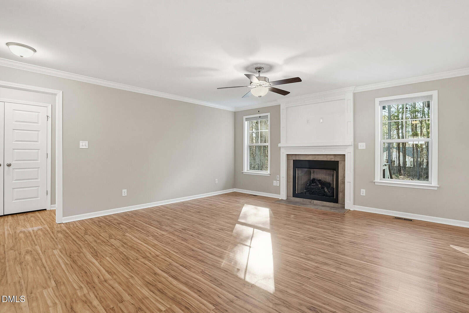 A spacious, empty living room with light beige walls, wood flooring, a ceiling fan with light, a white fireplace, two windows, and a white door. Natural light enters through the windows.