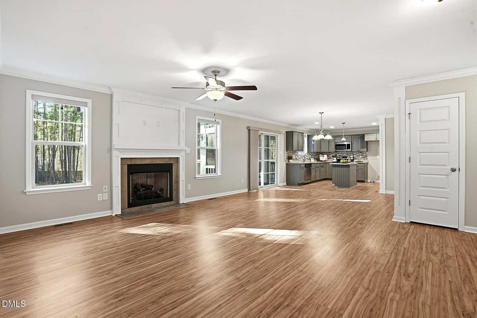 Spacious open-concept living area with light wood flooring, a fireplace, ceiling fan, and large windows; view extends to a modern kitchen with an island, stainless steel appliances, and dining area.