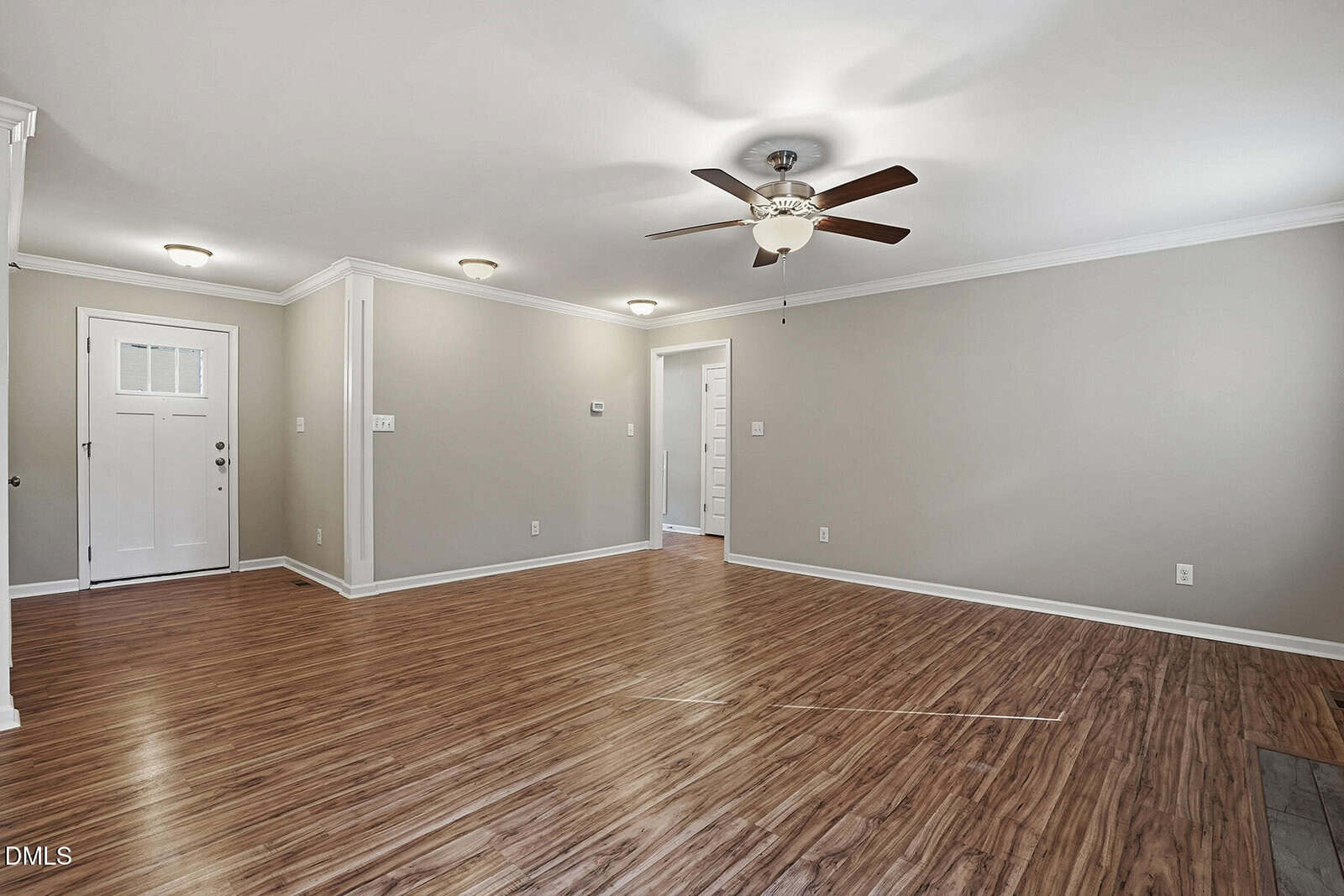 A spacious room with light wood flooring, beige walls, white trim, ceiling lights, and a ceiling fan. The room has multiple doorways, including a white entry door with glass panels.