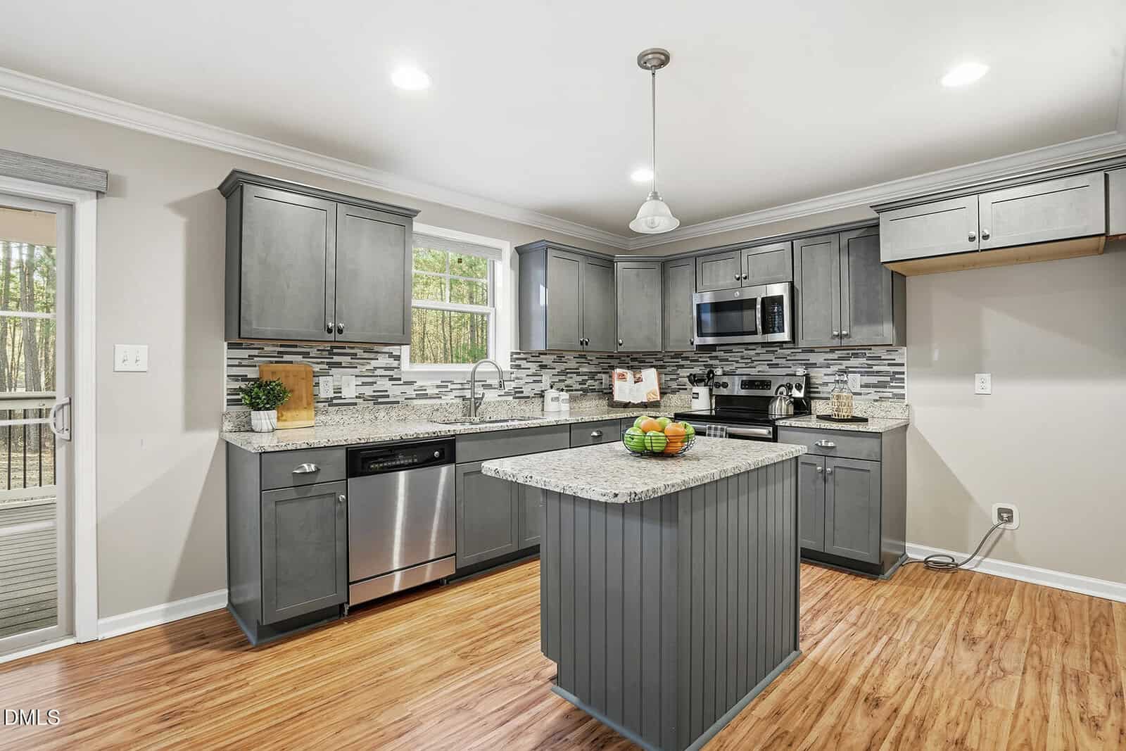 Modern kitchen with gray cabinets, stainless steel appliances, mosaic tile backsplash, and a small island with a fruit bowl. Hardwood floors and a window above the sink provide a bright, welcoming atmosphere.