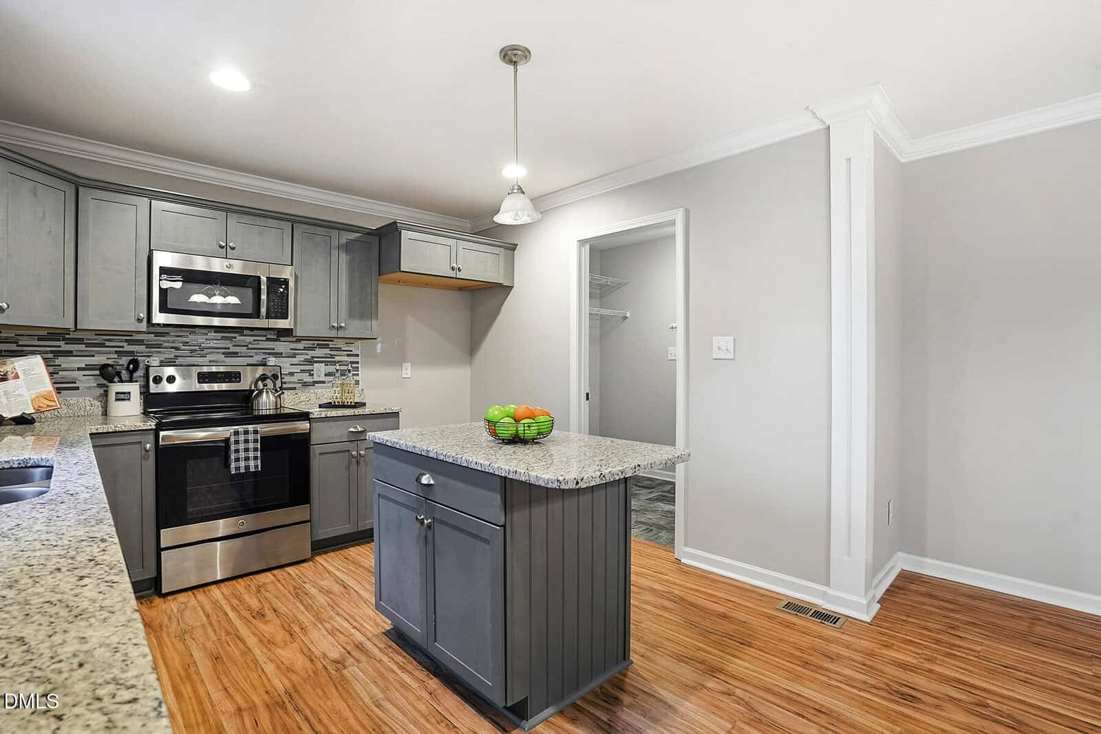 Modern kitchen with stainless steel appliances, gray cabinets, a small island topped with green apples, granite countertops, light wood floors, and gray tile backsplash. A doorway leads to a lit pantry area.