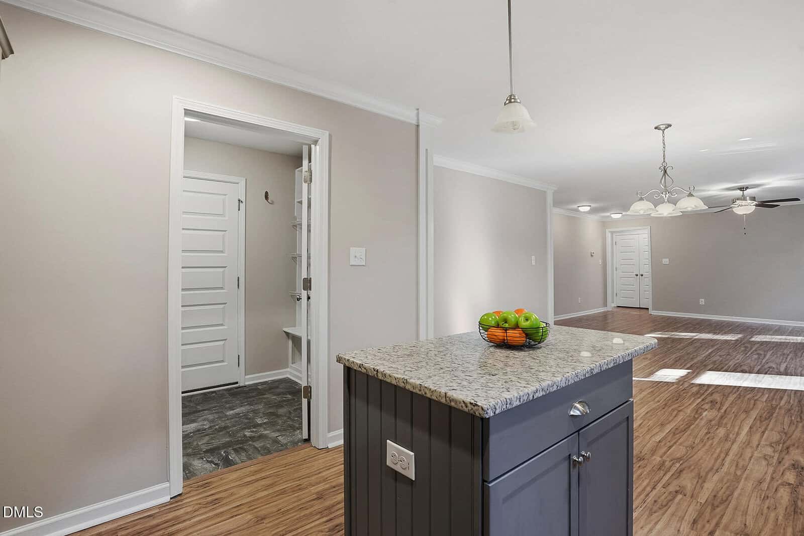 Kitchen island with a bowl of apples in the foreground, view of a bright open living space with wood floors, light-colored walls, and ceiling fans in the background.