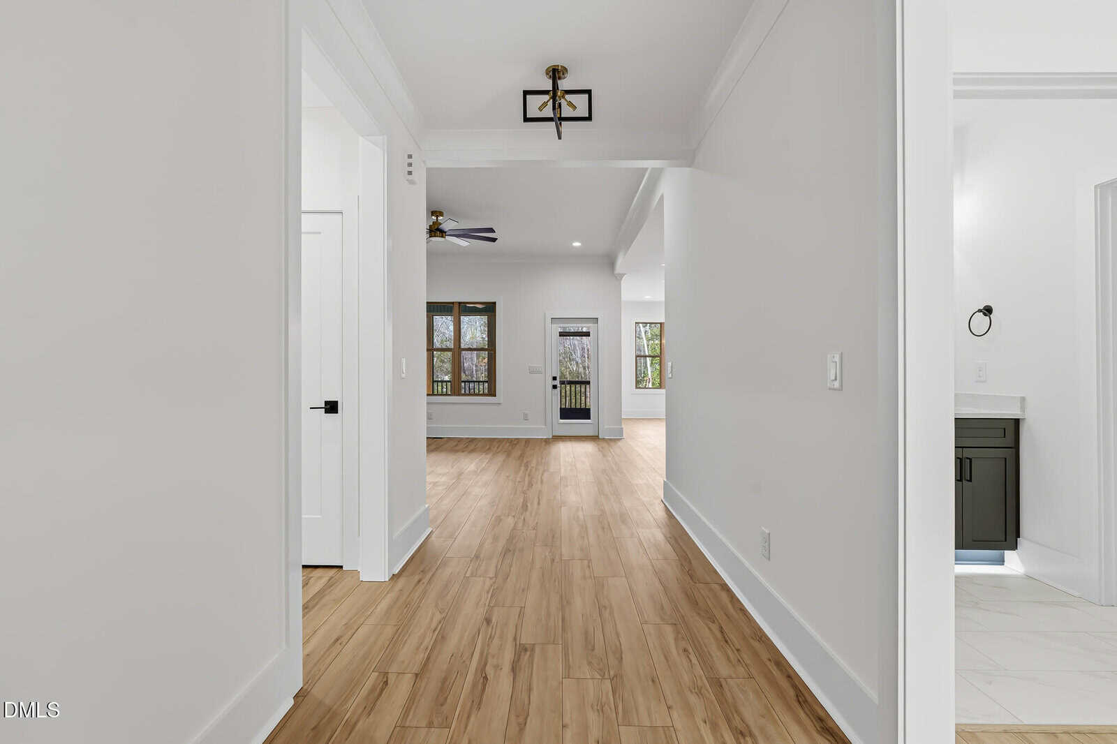 A bright hallway with light wood floors leads to an open living area with large windows and glass doors. White walls and modern fixtures create a clean, spacious feel. A partial view of a bathroom is visible on the right.