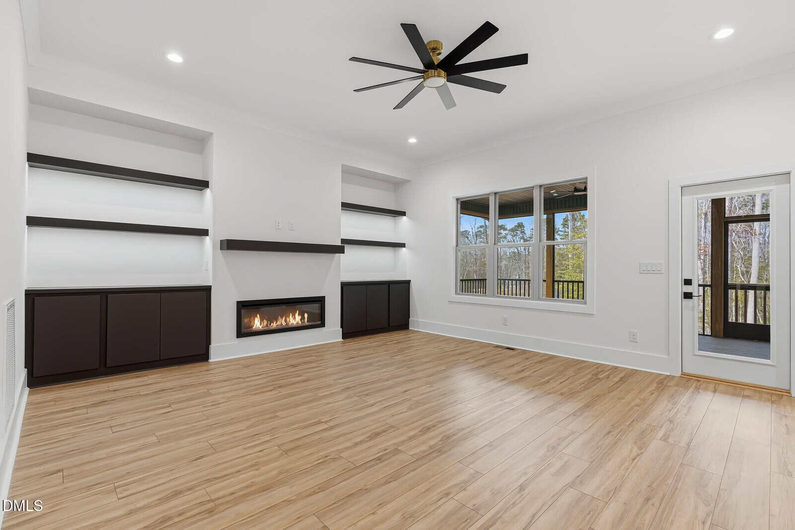 A modern living room with light wood floors, built-in shelves and cabinets, a sleek gas fireplace, a large ceiling fan, white walls, a window, and a glass door leading to a screened porch.