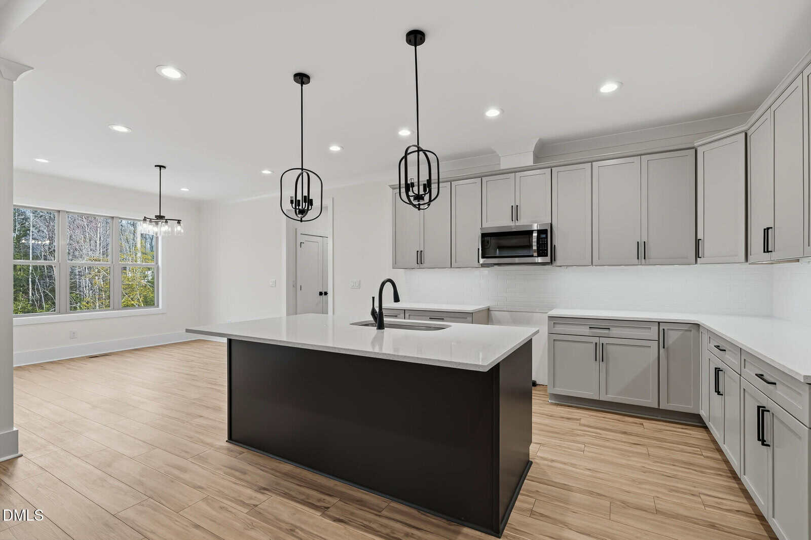 Modern kitchen with light gray cabinets, a large black island, white countertops, stainless steel appliances, pendant lights, wood-look flooring, and a dining area with large window letting in natural light.