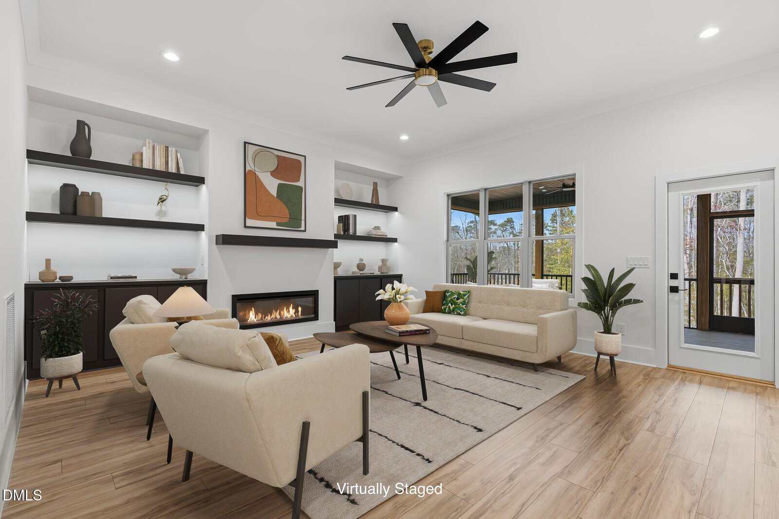 A modern living room with light wood flooring, beige sofas, a coffee table, and a fireplace. Built-in shelves with decor, a large abstract painting, and large windows brighten the space. The image is labeled "Virtually Staged.