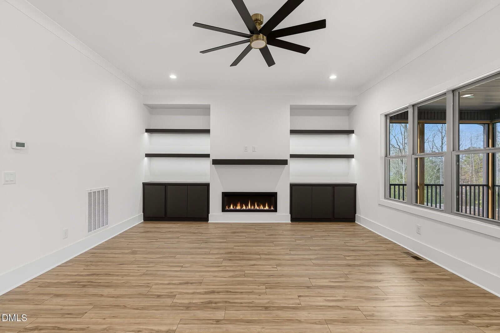 Modern living room with wood floors, white walls, built-in shelves, a central fireplace, black cabinets, a large ceiling fan, and a window overlooking trees.