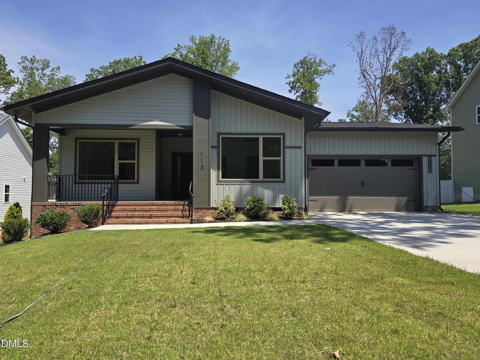 Single-story modern house at 113 Ottawa Drive, Louisburg, with gray siding, black trim, a covered front porch with railing, brick steps, large windows, a double garage, and a well-maintained green lawn under a blue sky.