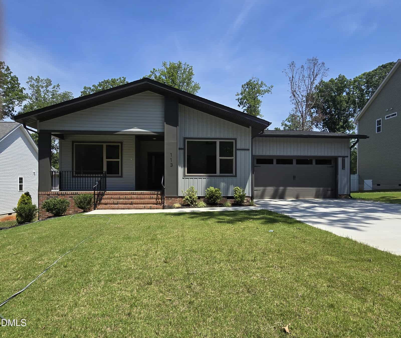 Modern single-story house with light gray siding, dark trim, a covered front porch, attached two-car garage, manicured lawn, small shrubs, and a paved driveway under a clear blue sky.