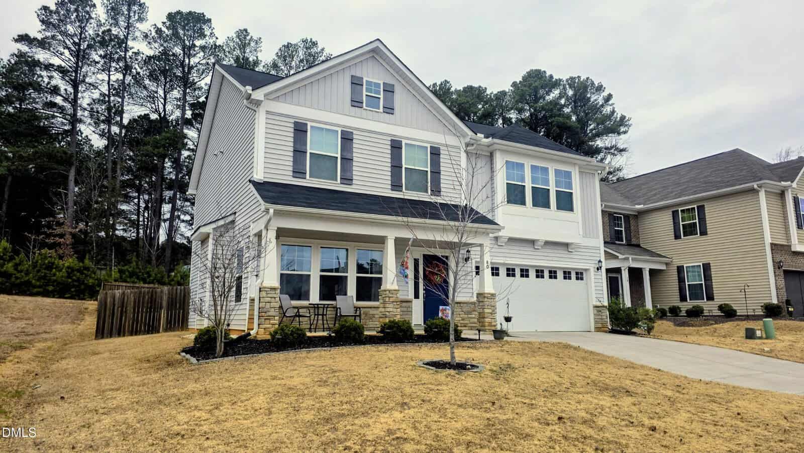 Two-story suburban house with grey siding, white trim, and stone accents, featuring a covered front porch, driveway, and garage. The lawn is brown, and trees are visible in the background.