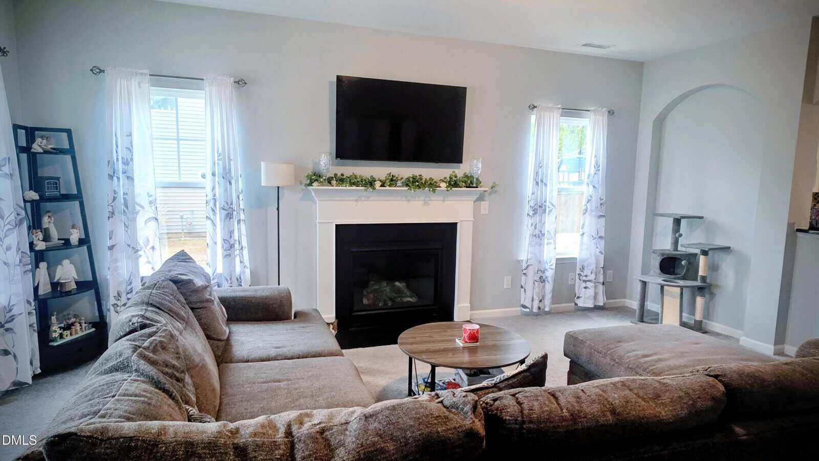 A cozy living room with a sectional sofa, round coffee table, fireplace with a garland, wall-mounted TV, two windows with sheer curtains, and a gray cat tree in the corner.