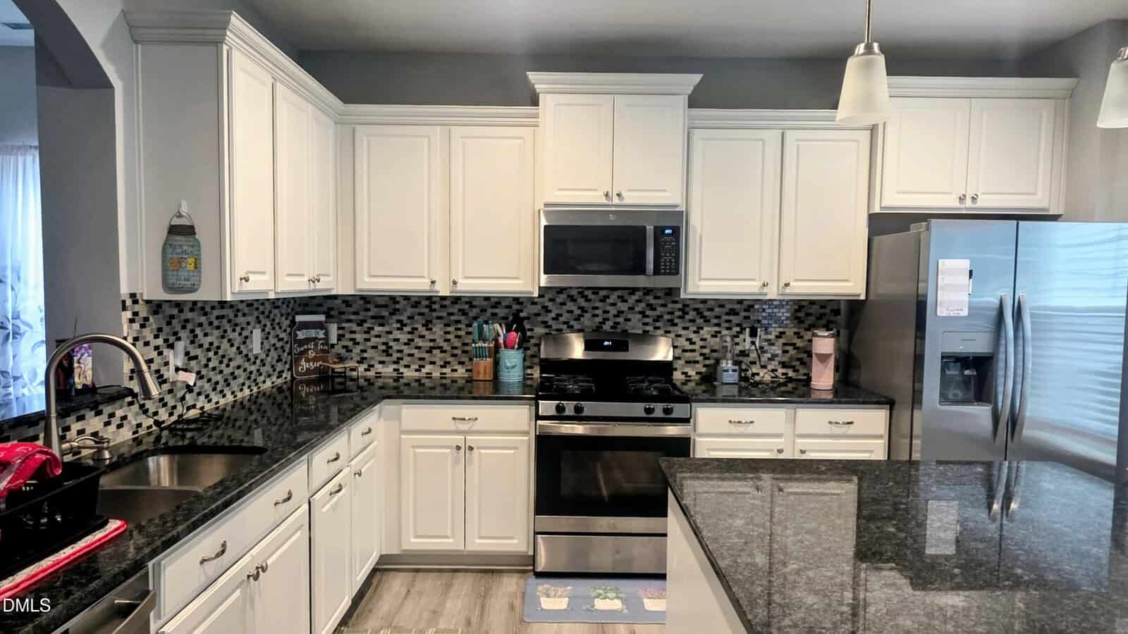 Modern kitchen with white cabinets, stainless steel appliances including a refrigerator and oven, black granite countertops, a mosaic tile backsplash, and pendant lighting over the counter.