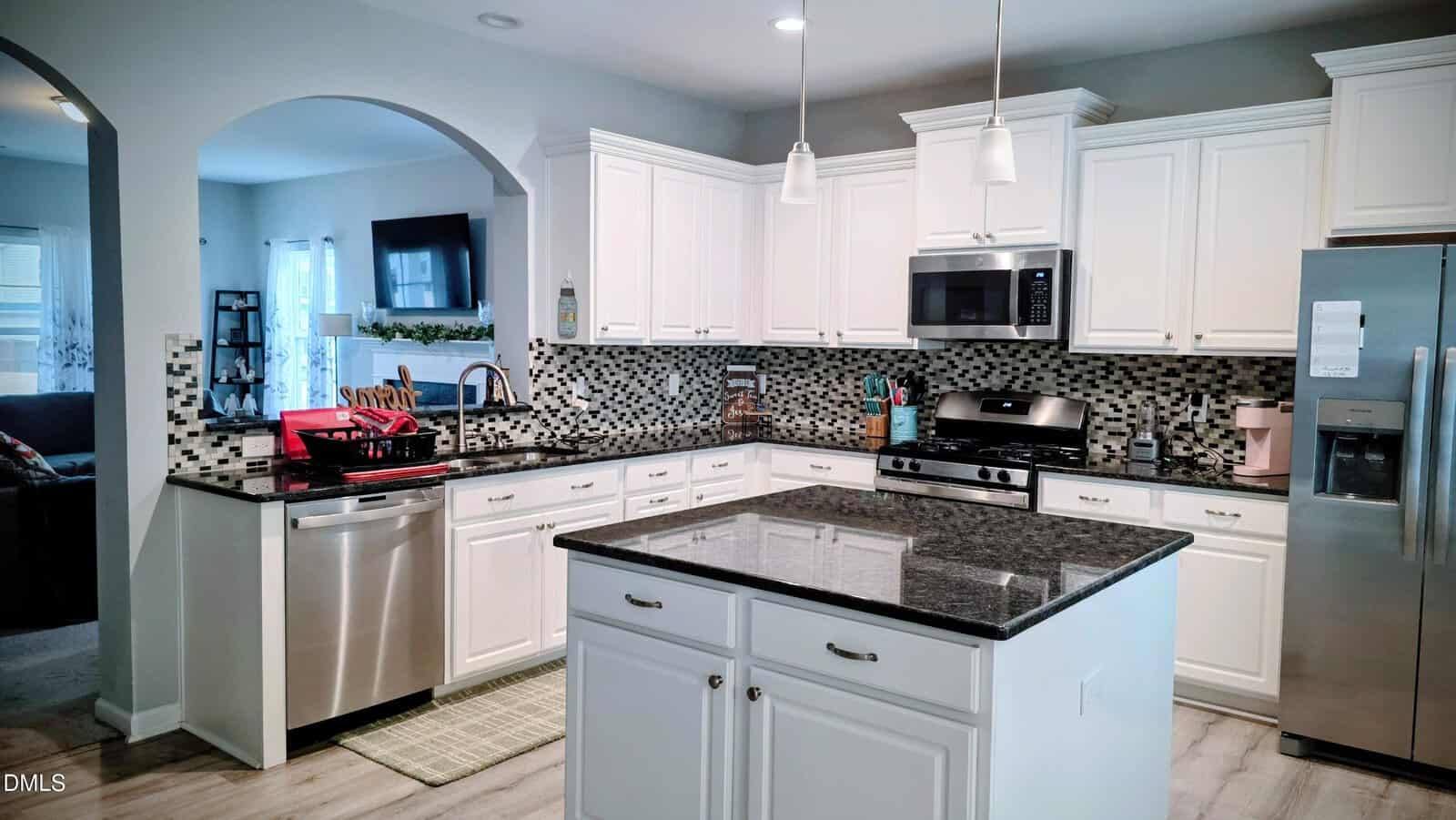 Modern kitchen with white cabinets, stainless steel appliances, mosaic tile backsplash, a central island with dark granite countertops, a dishwasher with a red dish rack, and pendant lights hanging from the ceiling.