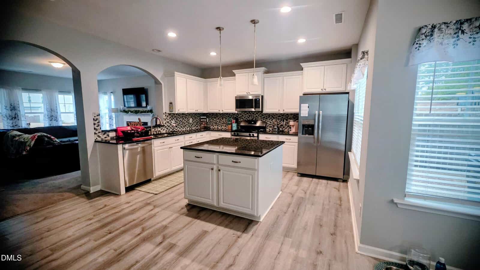 Modern kitchen with white cabinets, stainless steel appliances, a central island with a dark countertop, mosaic tile backsplash, pendant lights, and wood-look flooring. Bright windows provide natural light. Adjacent living area is visible.