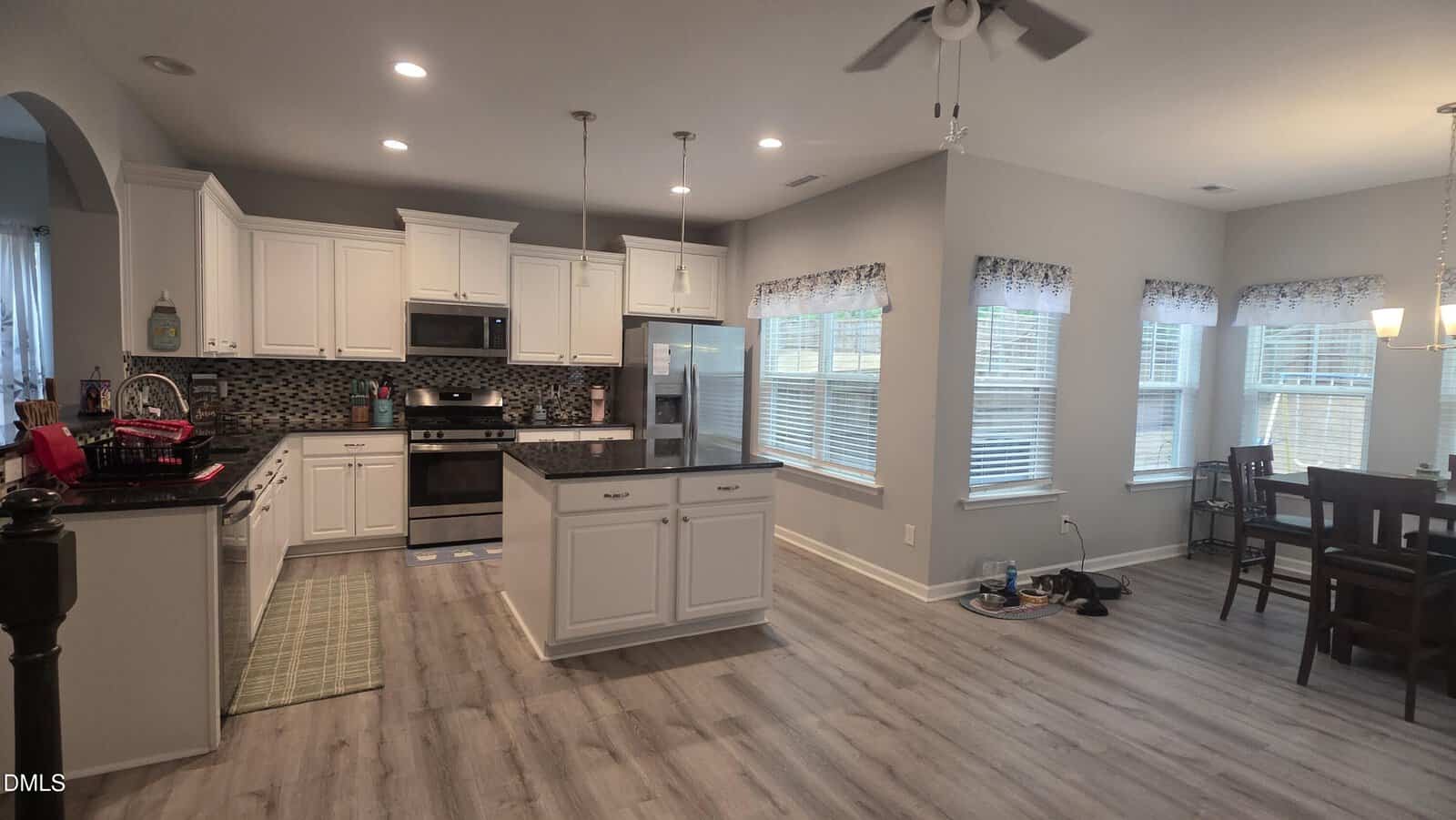 Modern, open-concept kitchen and dining area with white cabinets, stainless steel appliances, an island, light wood flooring, and multiple large windows letting in natural light. Dining table and ceiling fans are visible.