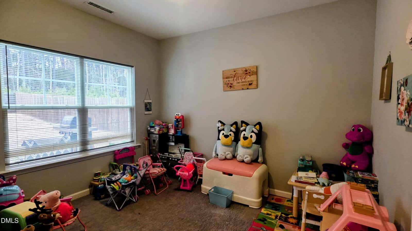 A children's playroom with large windows, filled with various toys and stuffed animals. Three plush Bluey characters sit on a chest, and a purple Barney toy is on a shelf. The room is bright and organized.