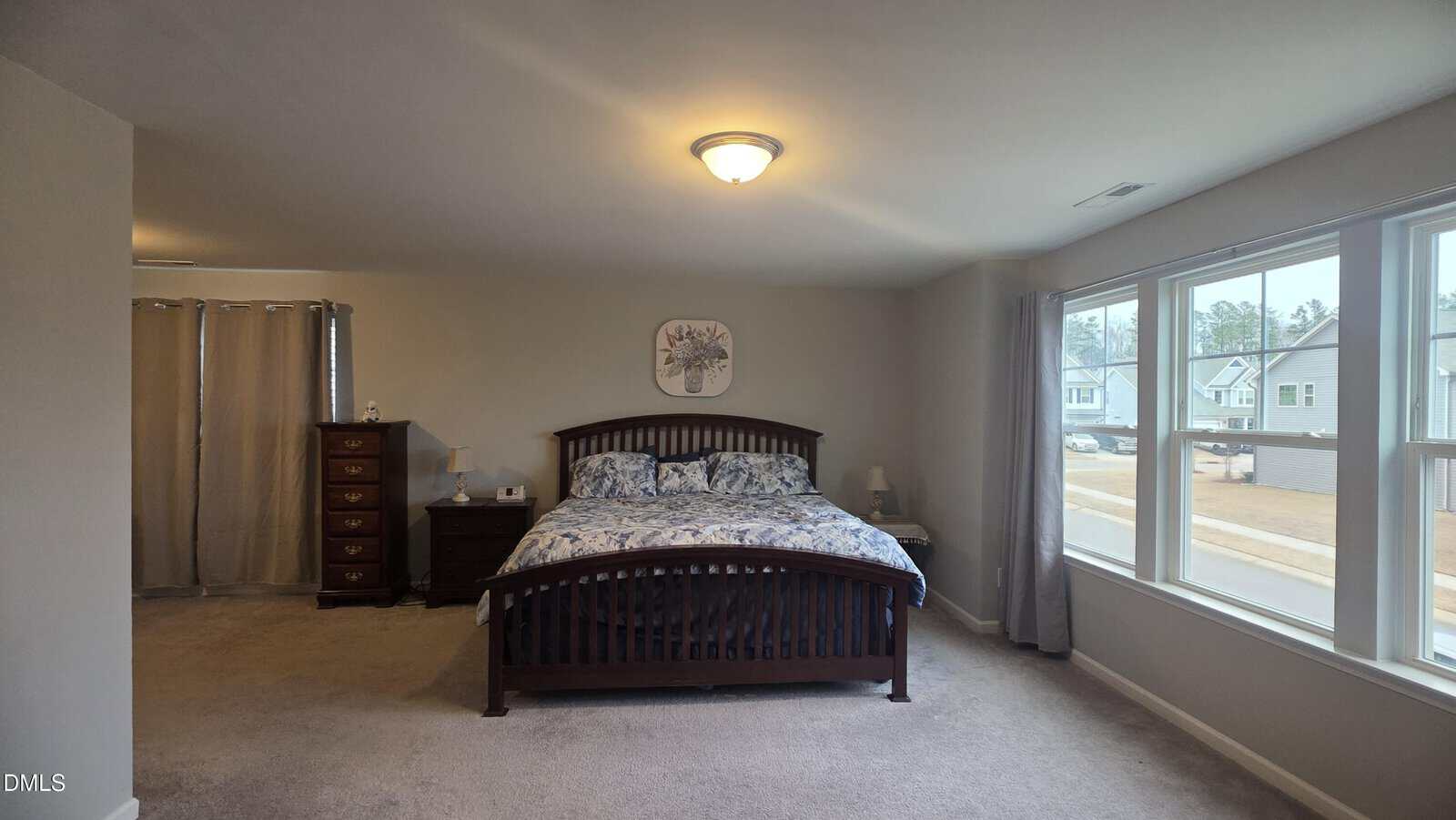 A spacious bedroom with a dark wooden bed, blue and white bedding, nightstand, tall dresser, carpeted floor, beige walls, and large windows letting in natural light.
