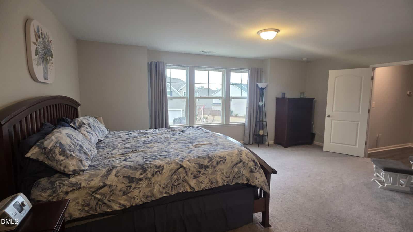 A spacious bedroom with a large bed, patterned bedding, dark wood headboard, dresser, and nightstand. Light beige walls, carpeted floor, and a window with curtains letting in natural light. Door opens to another room.