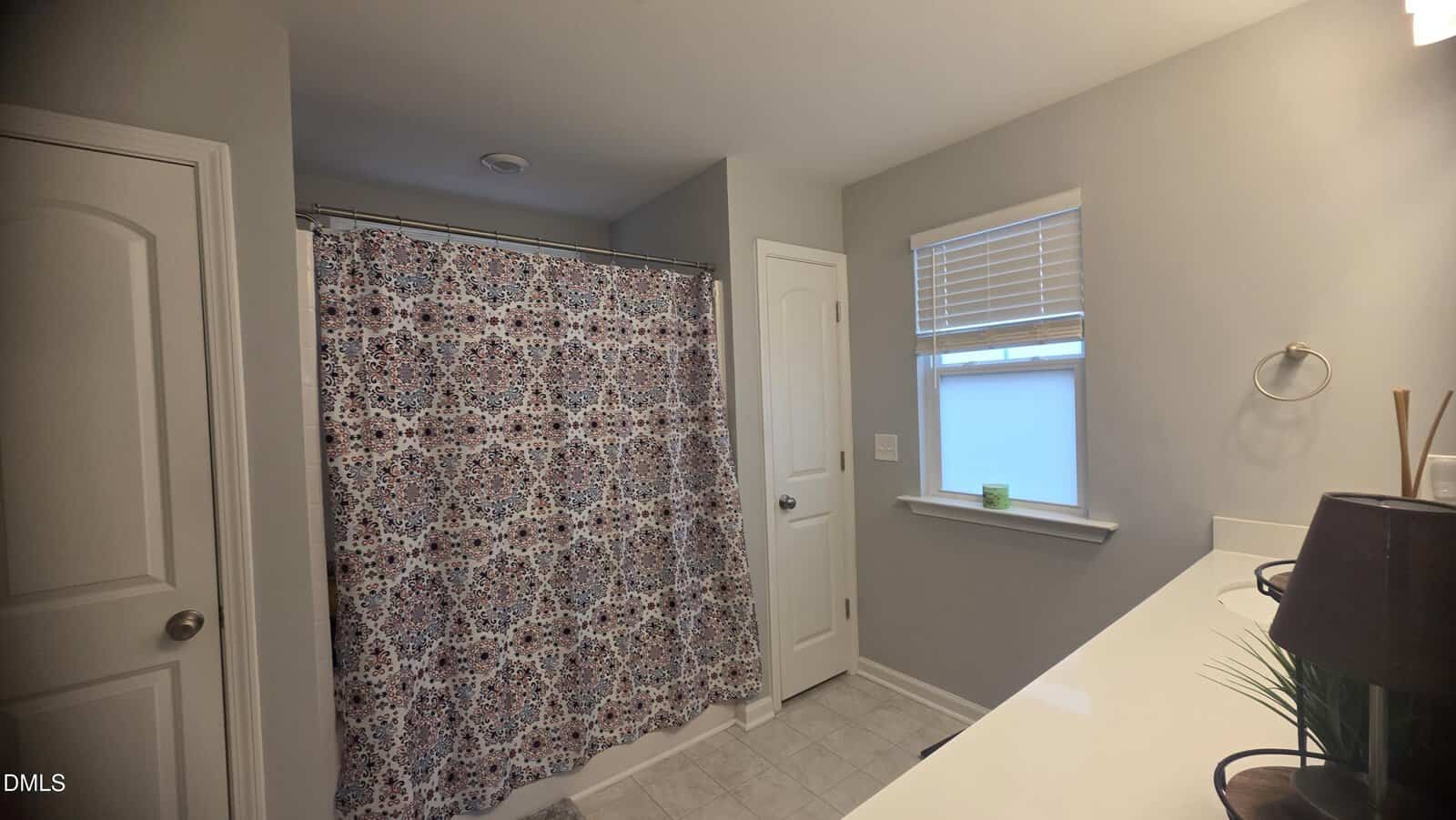 A bathroom with a patterned shower curtain, gray walls, white door, tiled floor, a window with blinds, a counter with a lamp, plant, and a green candle on the windowsill.