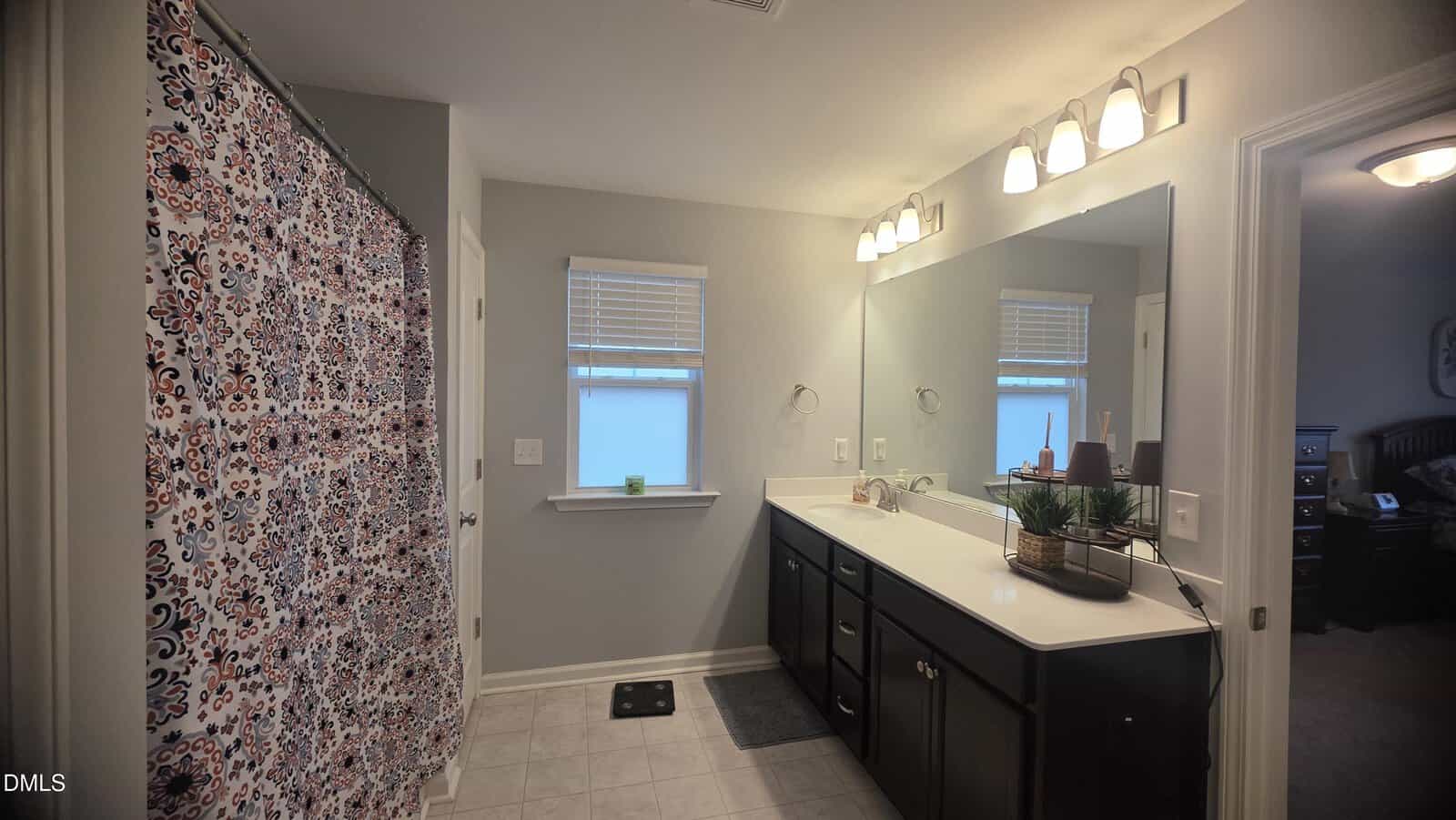 Bathroom with double sink vanity, large mirror, light fixtures above, patterned shower curtain on the left, tiled floor, two windows with blinds, and a doorway leading to another room.
