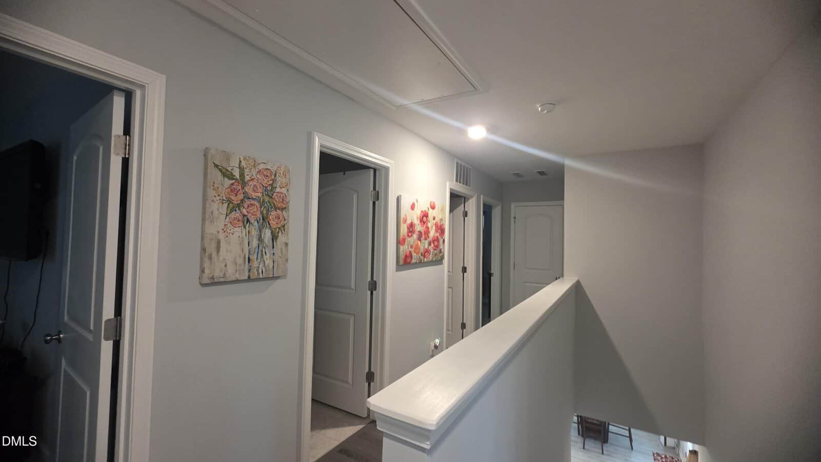 A hallway with light gray walls, three closed white doors, floral paintings on the walls, and a white railing overlooking stairs with natural light coming from below.
