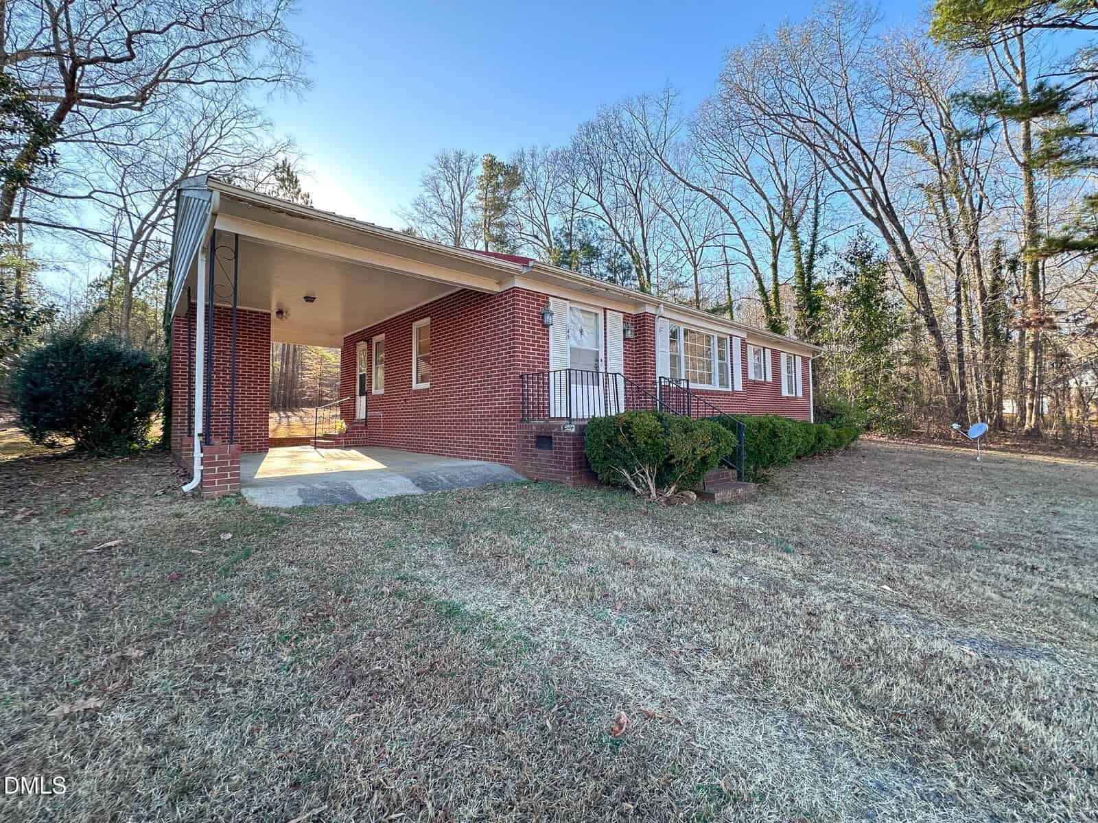 Single-story red brick house with a covered carport, white trim, front steps, and a grassy yard, surrounded by tall leafless trees under a clear blue sky.