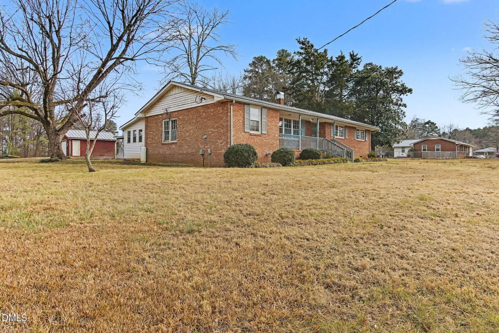 Single-story brick house with large front yard, shrubs near the foundation, leafless trees, and a clear blue sky. A detached garage and another house are visible in the background.