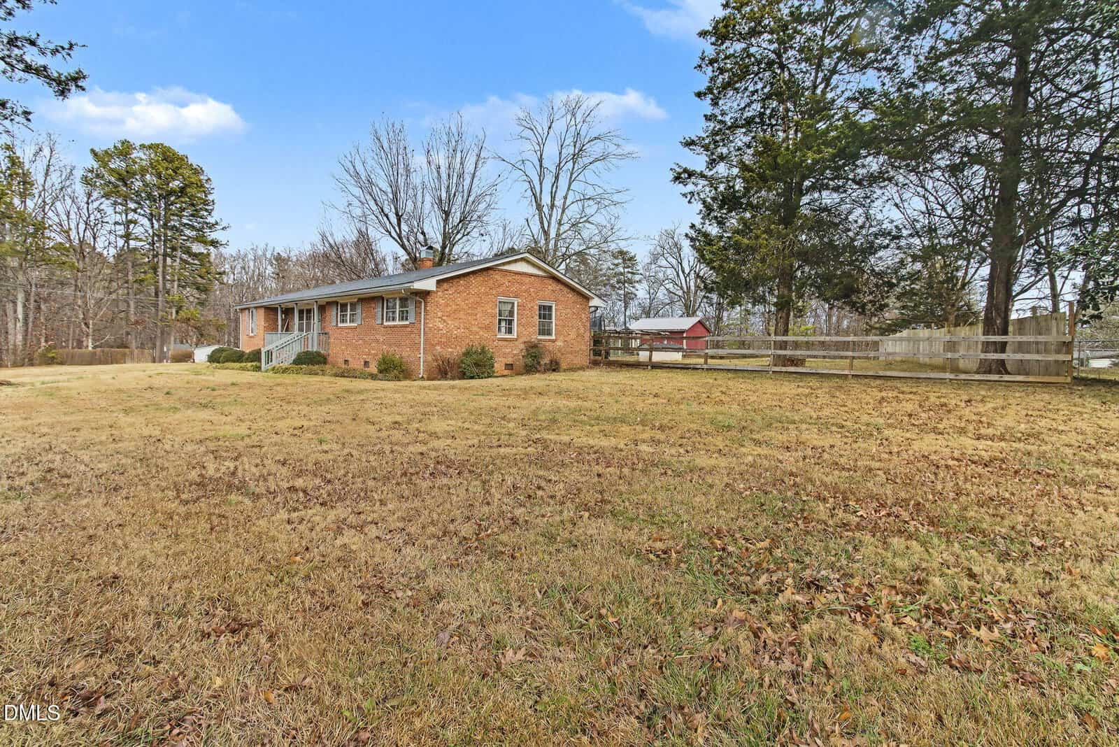 A single-story brick house with a large, grassy yard, surrounded by trees and bordered by a wooden fence under a blue sky with scattered clouds.