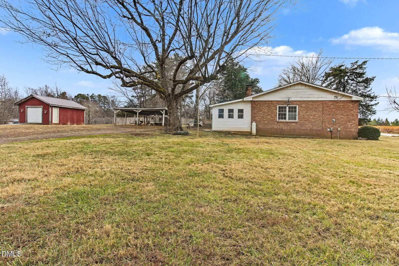 A brick house with white siding sits on a grassy yard with a large leafless tree. In the background, there is a red barn and a covered structure. The sky is partly cloudy.