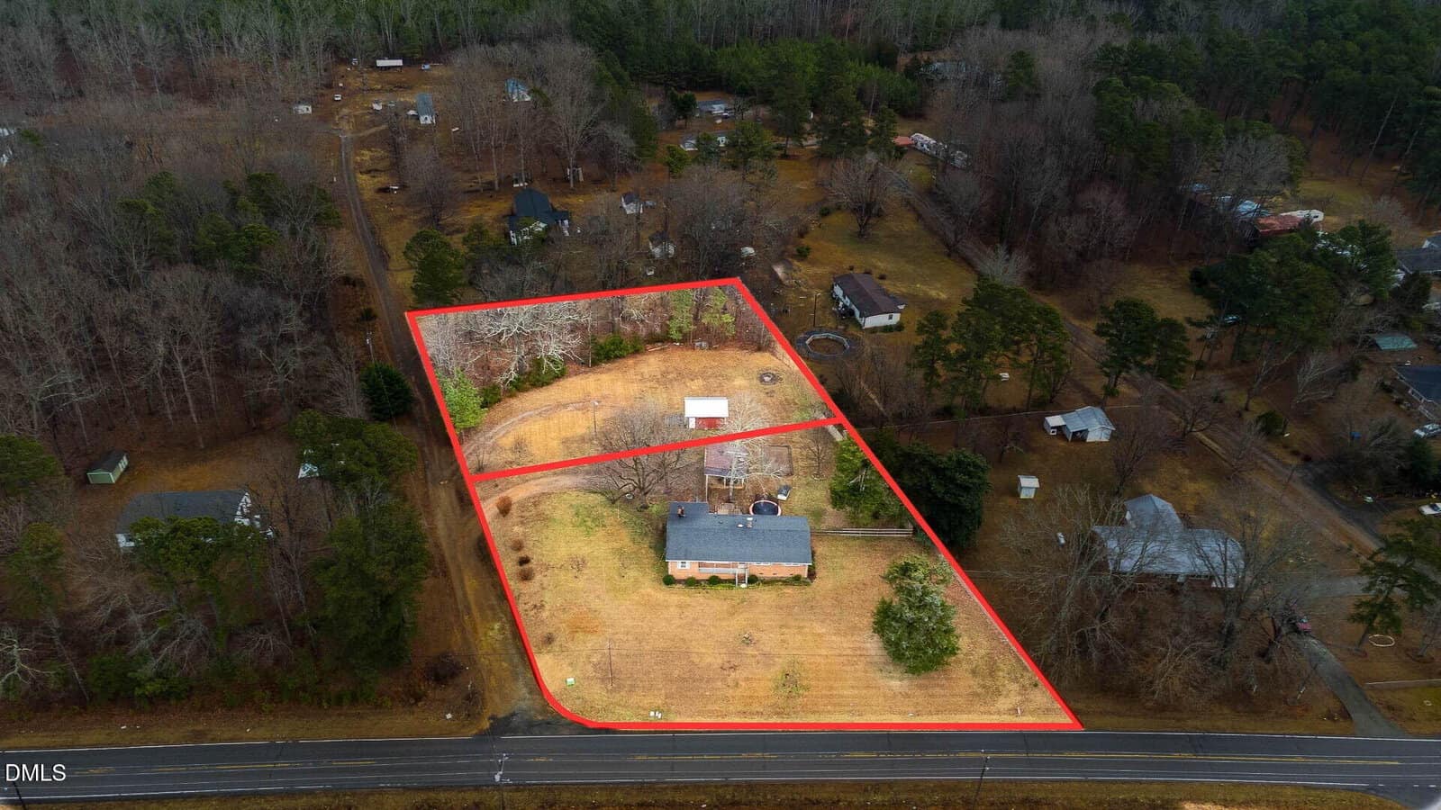 Aerial view of a rural property outlined in red, featuring a house, a smaller outbuilding, a driveway, and spacious yard, surrounded by trees and neighboring homes. The property borders a road in the foreground.