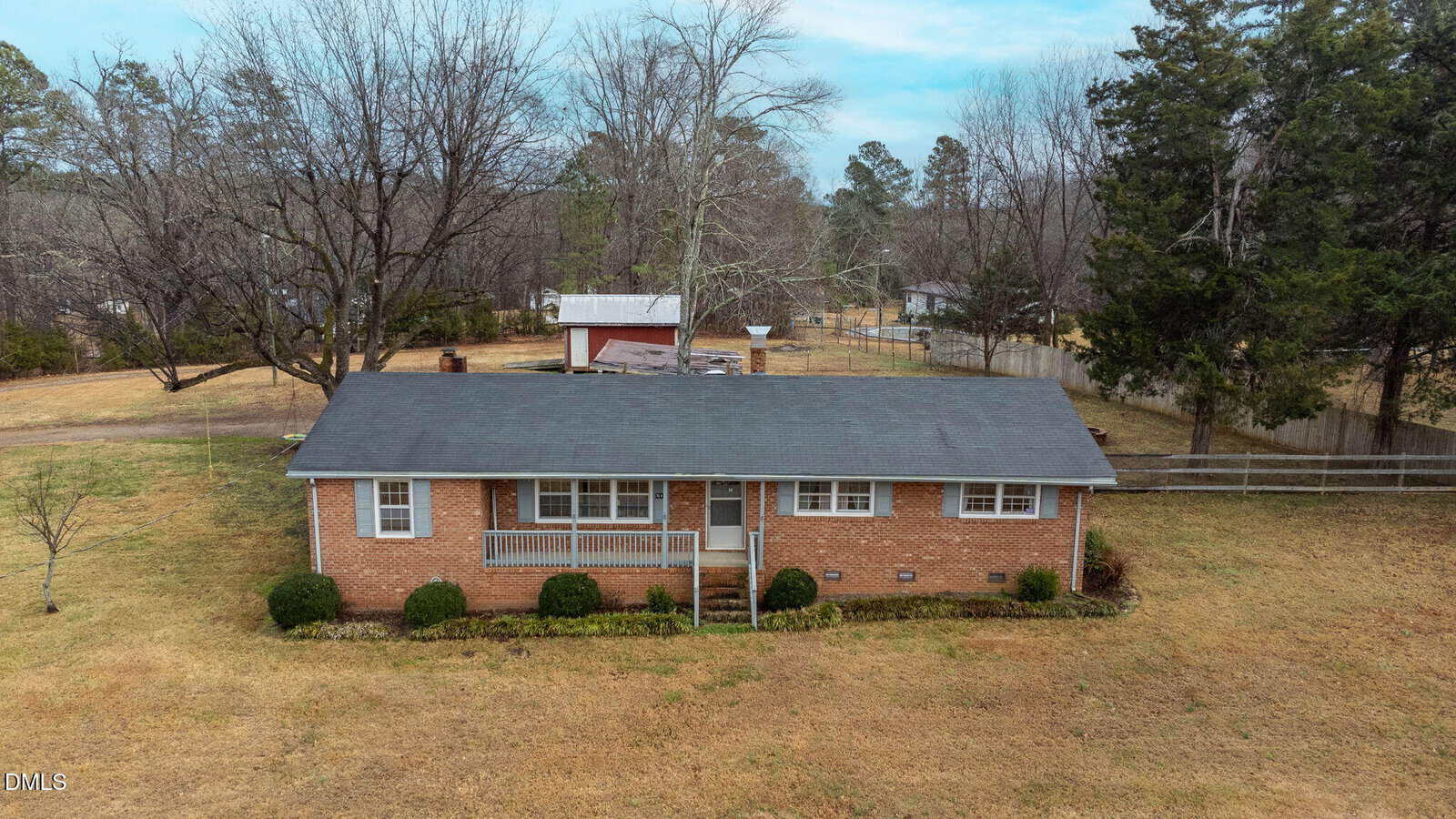 A single-story brick house with a dark roof and front porch, surrounded by a large yard with sparse grass and a few trees. A detached structure and more trees are visible in the background.