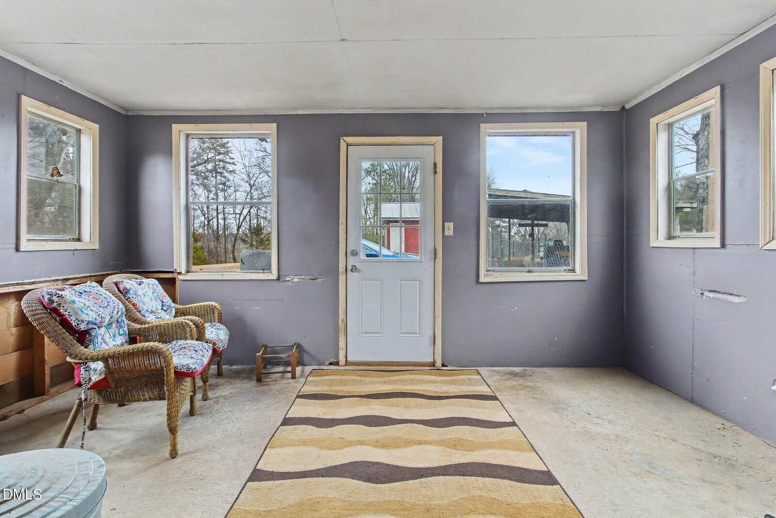 A simple sunroom with gray walls, five windows, a glass door, two wicker chairs with cushions, a patterned rug, and a pet food bowl on the concrete floor. Outdoor scenery is visible through the windows.