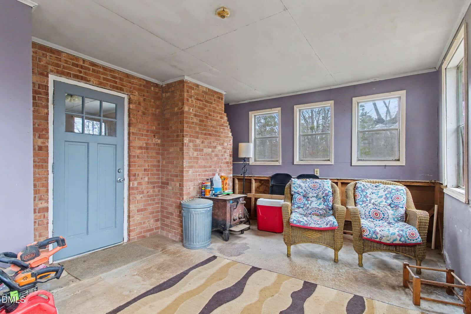 A small enclosed porch with a blue-gray door, brick wall, and purple accents. Two wicker chairs with patterned cushions, a rug, a lamp, coolers, tools, and cleaning supplies are visible. Several windows let in natural light.