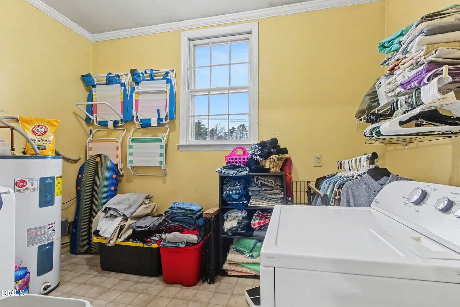 A laundry room with yellow walls, a washing machine and dryer, shelves of folded clothes, hanging shirts, an ironing board, laundry baskets, and stacked beach chairs next to a window.